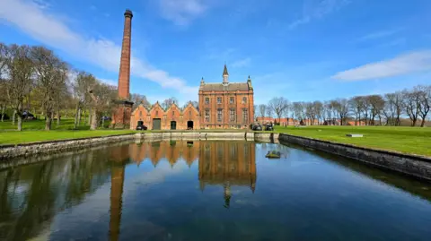View of a Victorian building - a former pumping station - in red brick with two high storeys and with tall windows, and a pitched roof with a small turret in the middle. To its side is a line of four brick buildings with pitched roofs and a separate high chimney. The scene is reflected in a body of water in front, with a line of leafless trees on either side under blue skies.