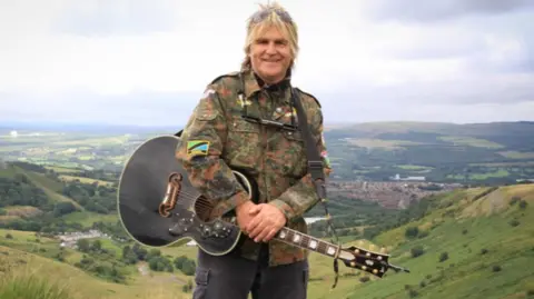 Mike stood in the countryside, you can see the hills behind him. He is holding a guitar with the strap around his shoulder and an army jacket on top. The man is The Alarm's Mike Peters