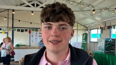 A young man wearing a pink polo shirt and a navy blue gilet smiles. He has wavy brown hair, blue eyes and has braces on his teeth.