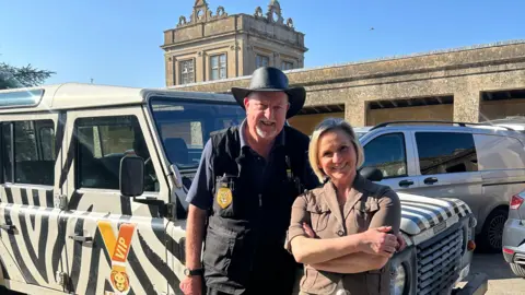 BBC Points West presenter Amanda Parr who has short blonde hair and is smiling is standing next to a man with a grey beard and a black cowboy-style hat. Behind them is a jeep-style vehicle with black and white stripes on it, and part of the main house at Longleat Safari Park