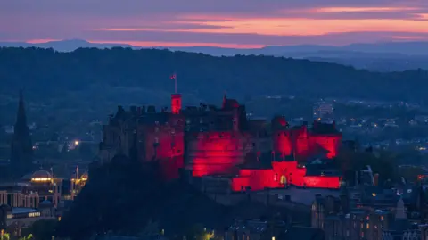 PA Media Edinburgh castle illuminated in red light against a dusk skye with the city houses in the background.