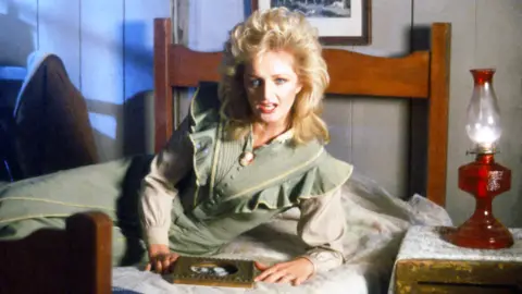 Getty Images Bonnie tyler in the video of the song "Holding out for a Hero" . She is lying on a bed in a sage green dress amd looking at a photo in a frame