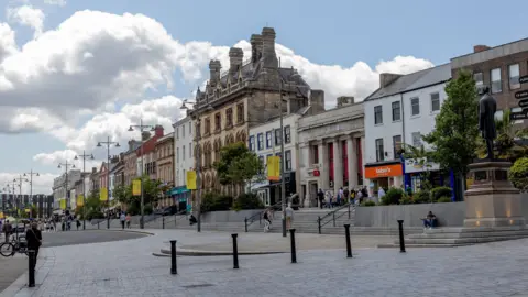 Wide view of a pedestrian-friendly town centre street lined with historic and modern buildings under a bright blue sky with large white clouds. Stone steps lead up to shops and businesses. A statue stands on a raised plinth at the right side of the square. Streetlights, trees, bollards, and scattered pedestrians are visible along the gently curving paved roadway and public plaza.
