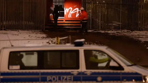 AFP via Getty Images Police and emergency personnel wearing uniforms and one a helmet inspect a metal box with graffiti on it beside a fence in the dark. A police van is seen in the foreground and snow is on the ground.