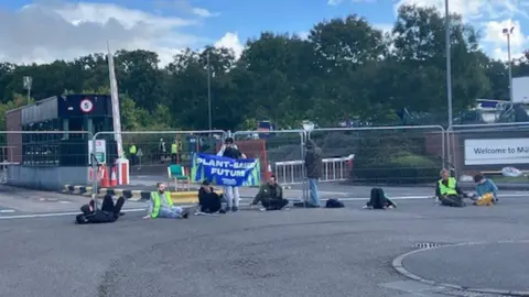 People are sitting or lying on the ground in front of a metal barricade. One of them is holding a banner reading: "PLANT-BASED FUTURE".