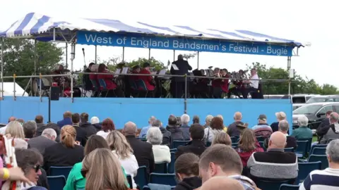 A brass band plays on a stage while a crowd watches on.