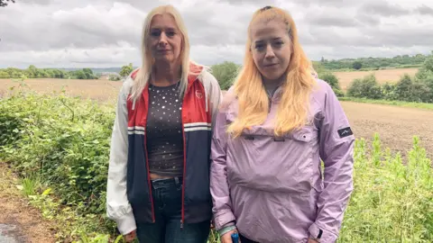 BBC/Lucy Ashton Two women standing in a field: The woman on the left has shoulder-length blonde hair and wears a red, white and blue sports jacket over a dark spotty top and jeans. The woman standing on her right has shoulder-length strawberry blonde hair and wears a lilac rain jacket.