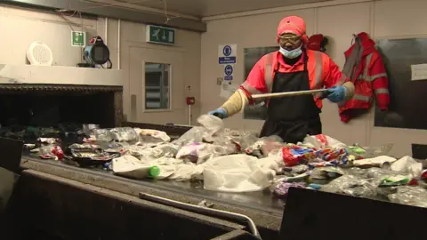 Inside a waste‑sorting facility, a worker in protective clothing stands beside a conveyor belt covered with assorted plastic items and other recyclables. The worker is using a long tool to sort through the materials. High‑visibility jackets hang on the wall behind, along with safety signs and equipment.