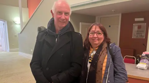 Paul Catling and Belinda Aldiss stand together in the foyer of Dereham Memorial Hall. He is taller than her, wearing a black coat. She has auburn hair and is wearing lots of layers under her blue jacket including a yellow jumper which is just visible. 