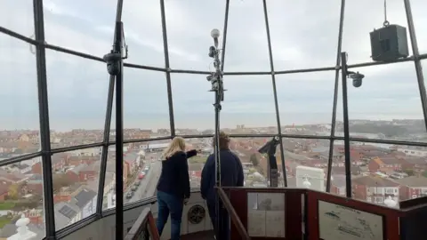 Two women with their backs to the camera in the lamp room of a lighthouse, high above the ground, with one pointing to a town below. The lamp room has three rows of big glass windows with black frames and metal posts with light bulbs on them. Towards the bottom of the picture are information displays. The town is made up mainly of red brick buildings. A road leads down towards the sea in the distance. The sky is grey.