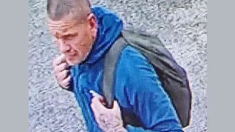 CCTV image of a man with short hair wearing a blue jacket with his hands tucked under the straps of a black rucksack.
