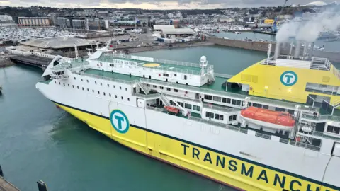 An aerial view of a white and yellow boat arriving into St Helier harbour. You can see rows of built up buildings and roads in the distance. Grey skies.