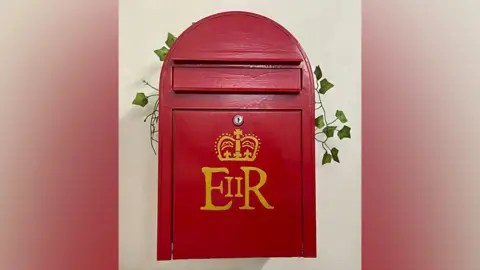 BAS/Aurelia Reichardt A wooden post box is painted red and fixed to a white wall. It has a gold crest and the initials ER on the front, indicating Queen Elizabeth II. A sprig of ivy is placed on top as decoration.