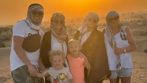 A photo of Hollie and her family dressed in holiday clothing. They are stood in a desert and there is an orange sunset behind them.