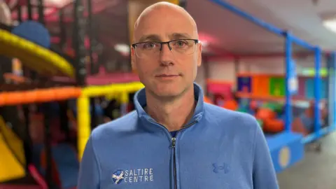 Craig Meikle wearing his Saltire Centre top. He is standing in front of  colourful soft play equipment (out of focus).