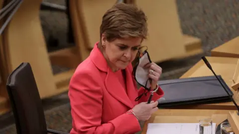 Nicola Sturgeon - a woman with short dark hair, wearing a pink jacket - pictured removing a face mask in the Scottish Parliament chamber during the pandemic