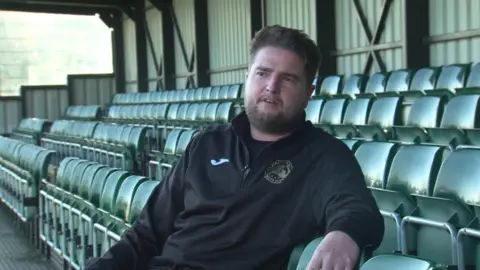 John Percival with a beard and brown hair sits in an empty stand of green seats talking to the camera.