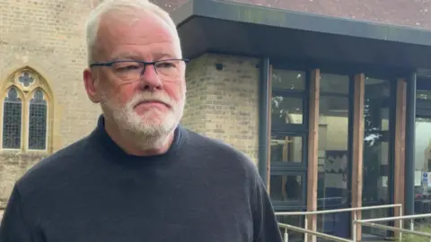 Peter is standing in front of the church hall that will host Broadstone Town Council meetings. He is wearing a dark grey jumper and glasses