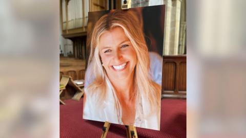 An image of a blonde woman smiling. The picture is on a large stand in a church. The carpet in the background is red.