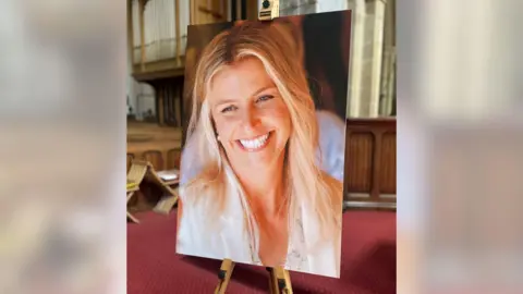 An image of a blonde woman smiling. The picture is on a large stand in a church. The carpet in the background is red.