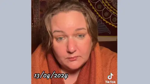 A TikTok freeze frame of Fiona Ryan, showing her looking at the camera as she makes a post. She is wearing an orange jumper