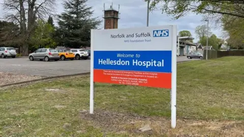 The picture is a close up of the sign for Hellesdon Hospital. It says Norfolk of and Suffolk NHS Foundation Trust is white red and blue. It also says "Welcome to Hellesdon Hospital". There is a car park behind the sign and some of the cars are visible. The sign it is positioned on grass. 