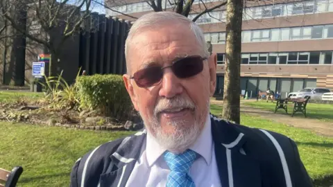 Chris Foote-Wood, wearing a striped blazer and blue tie along with dark glasses, smiles as he stands in a green park outside a public library. 