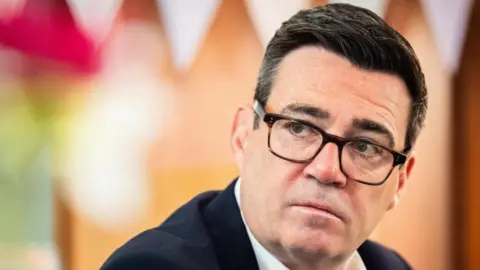 Prime Minister Andy Burnham wearing a dark suit and glasses. 