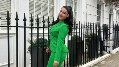 Nadia Suliaman Picture of Nadia Suliaman pictured in a bright green suit. She stands outside black fencing around a white townhouse. She smiles with her head titled to the side at the camera. She has very dark brown hair, pulled half up and half down. She has brown eyes. 