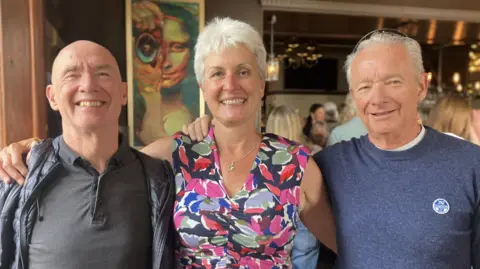 A woman with short white hair wearing a floral dress stands between two men. The man on the left is wearing a black shirt and the man on the right is wearing a blue jumper. They are all smiling at the camera and have their arms around each other's shoulders.
