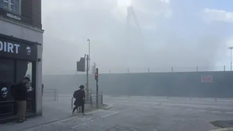 Supplied A person wearing a dark jacket and hat walks across the road as a haze of dust lingers in the air obscuring the demolition site behind it