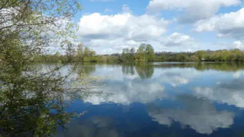 North Northamptonshire Council A large expanse of open water, with trees on the far bank and in the foreground on the left. White clouds and blue sky are reflected in the water.