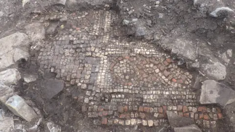 A photograph of an exposed mosaic floor. The mosaic sits in the centre of the image with red and white bricks in a spiral pattern. Around it you can see the excavations which have taken place and on the left hand side stones which look like the base of a wall.