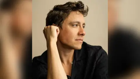 Jude Guaitamacchi, a man wearing a black shirt, is looking to his left while propping his head up on is arm. He has brown hair and is featured posing against a light coloured backdrop.