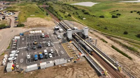 A drone shot of a car park that has a few cars parked, a rail line with a stationary train on the tracks and a footpath over the railway line, and on the right hand side of the train track is green fields with a small lake in the distance. 