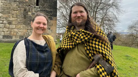 Caroline Nicolay and Thomas Timbrell - a pair of living history reenactors dressed in Iron Age clothes - standing in front of a castle. Thomas has an iron age axe in one hand.