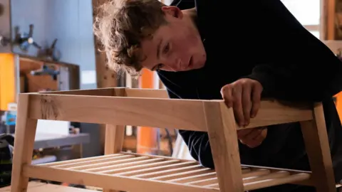 Elis working on a piece of wooden furniture. He is bent over as he focuses on his work and wears a black hoodie.