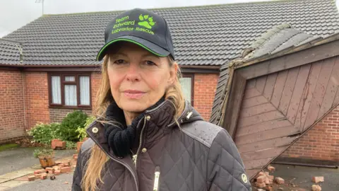 Daughter of the homeowner, Wendy Brassington. She is wearing a cap and a brown coat. She is stood in front of a car port which has been knocked down.