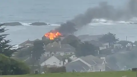 A photo taken from a field overlooking a large house filled with flames with black smoke pouring out. There are houses all around it and the sea beyond it.