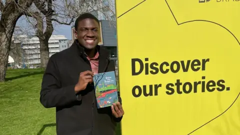University of Bradford/David Ben Eke A man smiles at the camera while holding a book. He is wearing a dark wool black coat and red shirt. He is standing outside next to a yellow sign that reads 'discover our stories'