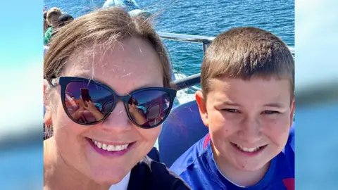 Julie Blackburn A woman with tied-back long brown hair wearing sunglasses sits on a boat next to a boy wearing a blue and red T-shirt. 