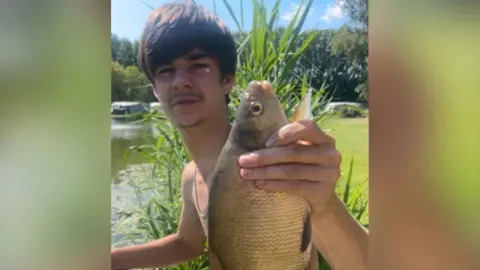 Holton Knott looking at the camera holding up a fish. He is sitting next to a riverbank.
