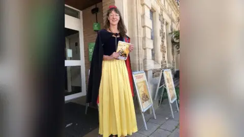 Sophia Moseley Ms Moseley pictured outside the theatre dressed as Snow White while holding a theatre programme. She has long brown hair and is wearing a yellow floor-length skirt with a navy blue top and navy blue cape.