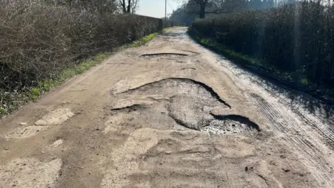 A view of lower Aldercar Lane which has not had any repairs made yet. Large cavernous potholes are visible in the foreground which take up nearly the entire width of the road.