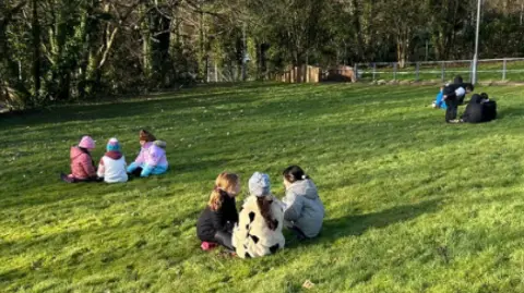 Simone Beach Children in groups in their coats sitting on the grass.