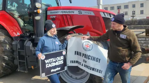 The picture shows Victoria Broughton and Nigel Witcombe standing in front of a red tractor on Weston-super-Mare's seafront.   They are holding two banners. One banner says "With our farmers", the other says "Farmers to Action"