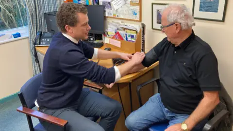 A man wearing blue jeans and a black top is having his blood pressure checked by a doctor. The doctor is sat on a chair wearing navy trousers and a navy jumper. They are sat in a small surgery room.