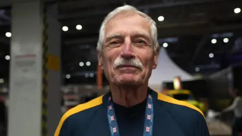 Roger Stainforth has short, neat grey hair and a moustache. He is standing in a gym and is wearing his medal around his neck. 