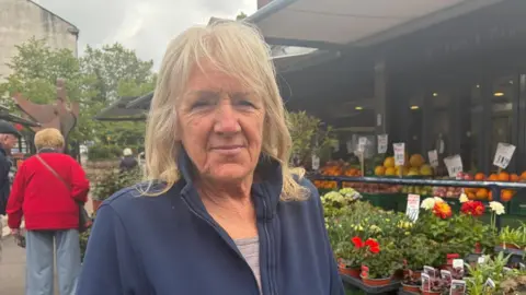 A woman with blonde hair and wearing a navy jumper stands in front of an outdoor flower and fruit market stall.