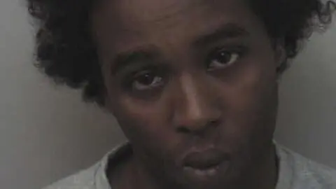 Fuad Awale, in a grey sweatshirt and with black afro-style hair, looks at the camera with his head tilted to the right as police take a mugshot 